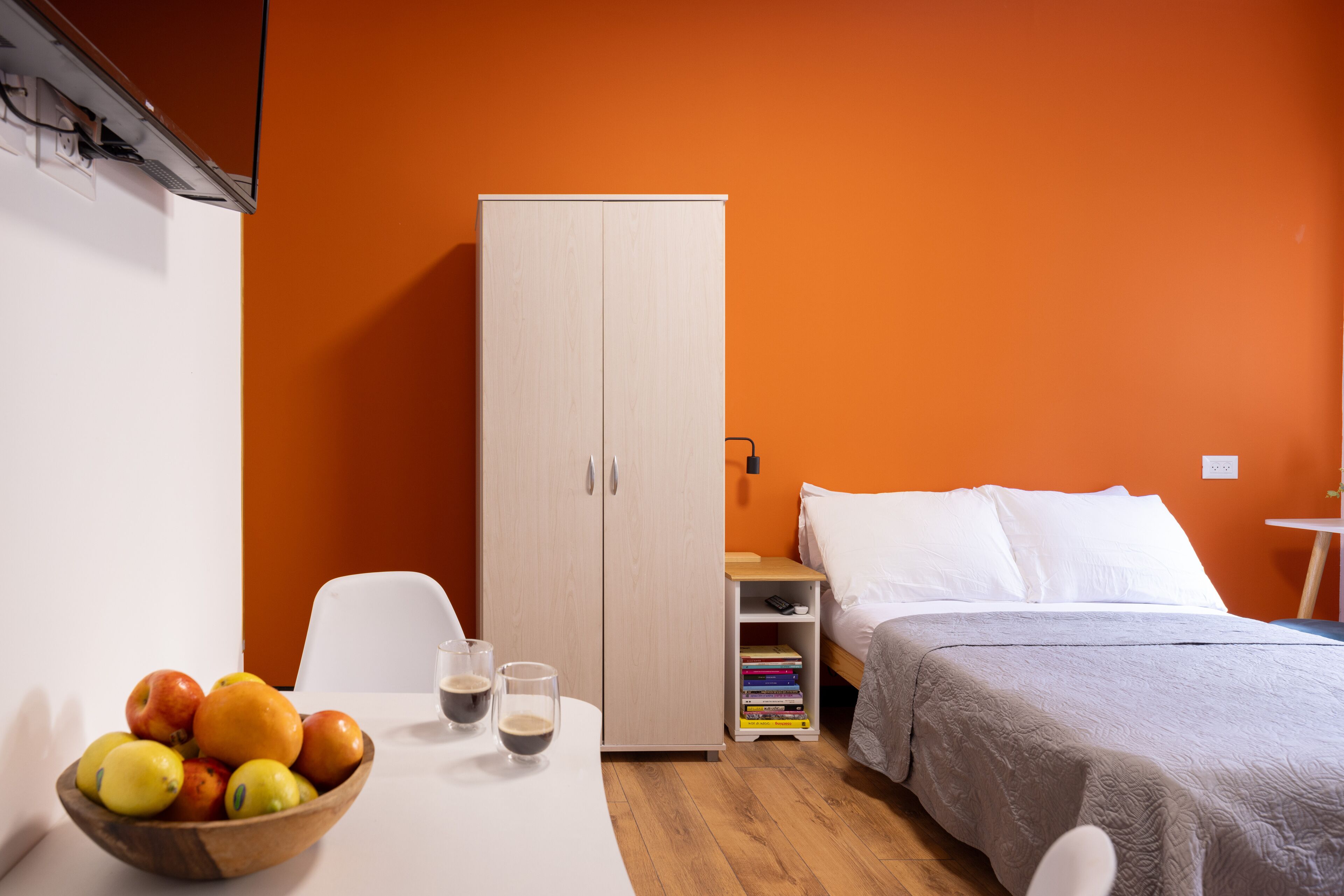 A room with a bed, a wardrobe, a table with fruit and drinks, and a bedside table with books and a lamp.