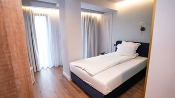 A bedroom with a bed, wooden flooring, white curtains, and a wall-mounted light fixture.
