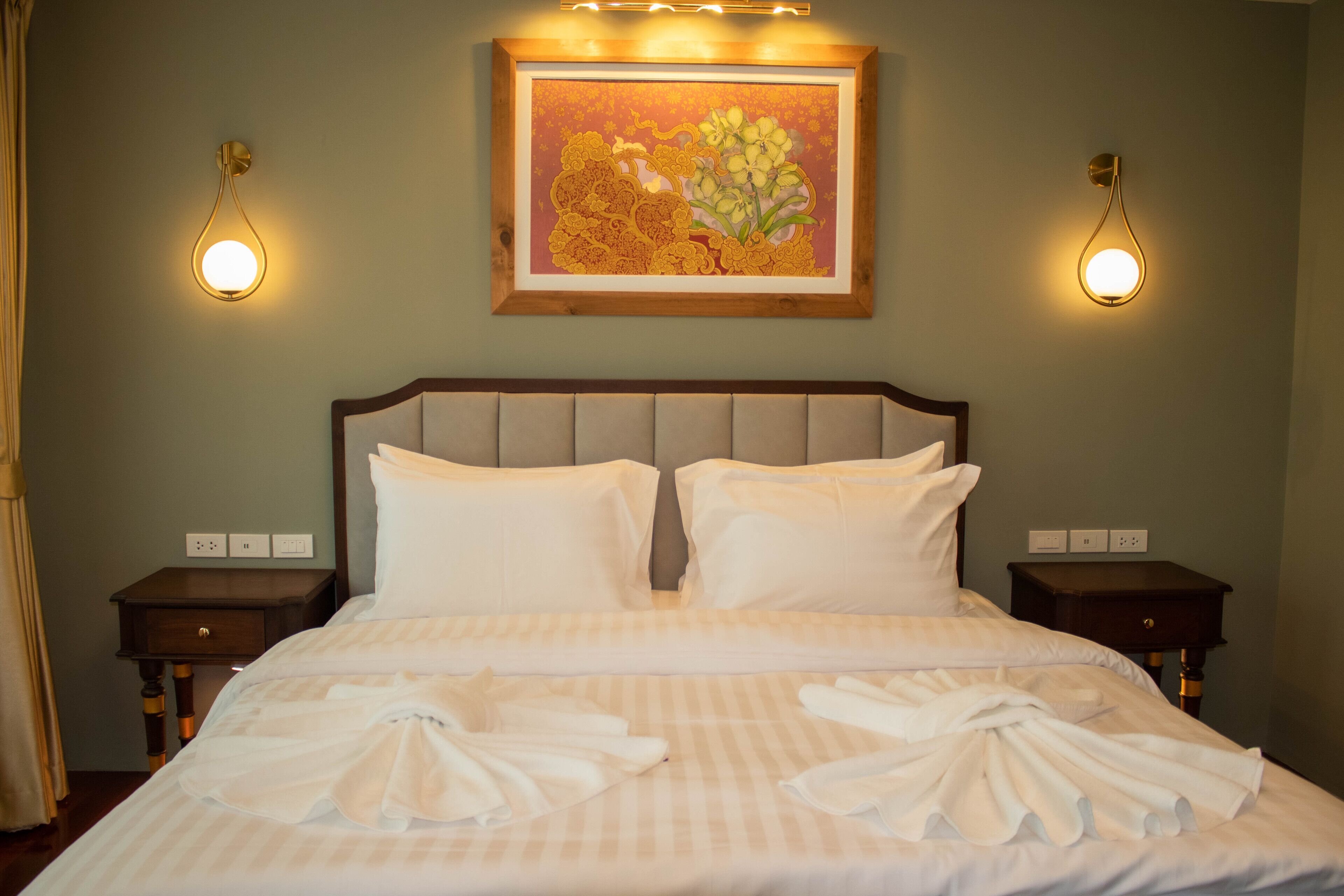 Ein ordentlich bezogenes Bett mit weißen Leinen und Handtüchern, ein gerahmtes Blumenbild darüber und zwei Nachttische mit Wandleuchten.