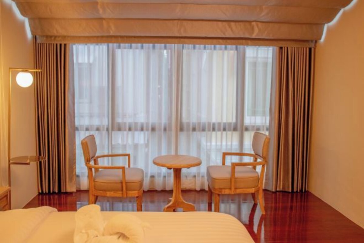 Hotelkamer met een bed, twee fauteuils, een klein rond tafeltje en een groot raam met gordijnen.
