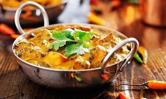 Un recipiente metálico tradicional con asa, probablemente conteniendo curry, adornado con una hoja, sobre una superficie de madera.