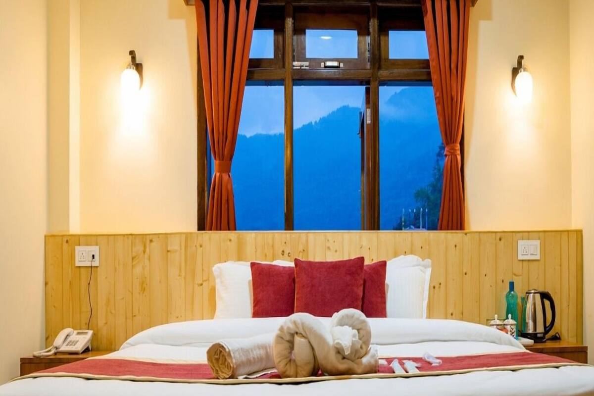 Habitación de hotel con cabecera de madera, cama grande con ropa de cama blanca y roja, vistas a las montañas a través de una ventana con cortinas y una mesita de noche con teléfono y hervidor.