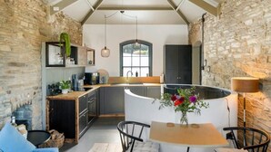A cozy kitchen with stone walls, a wooden dining table, and a built-in island with a sink. - Castle by the Beach with Sea Views, Portreath (Redruth)