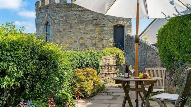 A stone building with a round tower, a table with chairs, and an umbrella. - Castle by the Beach with Sea Views, Portreath (Redruth)