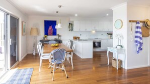 A modern kitchen with white cabinets, a central island, and a dining area with white chairs and a blue and white patterned chair. - Bimbajong Main Beach - Pet Friendly- Oceanstays (Yamba)