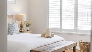 A bedroom with a bed, a bedside table with a lamp, a window with blinds, and a wooden bench. - Bimbajong Main Beach - Pet Friendly- Oceanstays (Yamba)