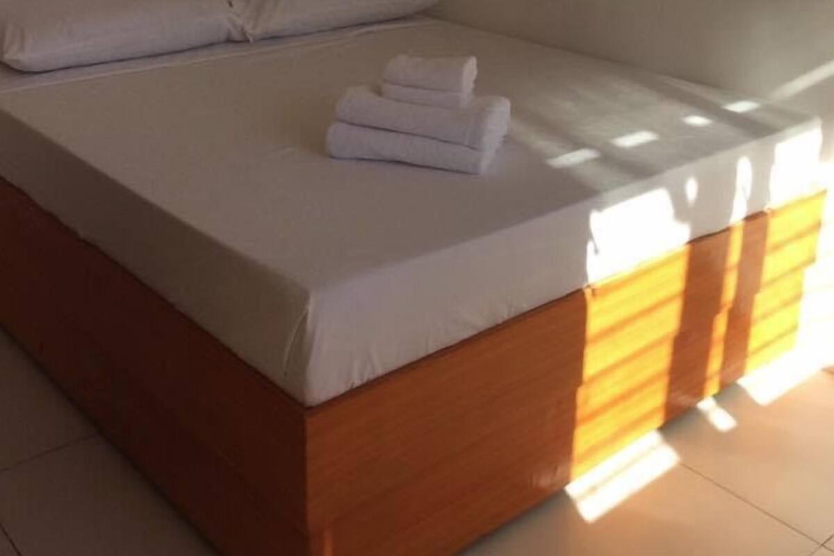 Un lit simple avec du linge de lit blanc et une tête de lit en bois, une serviette pliée posée sur le lit, et une fenêtre qui laisse entrer la lumière dans la pièce.