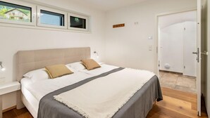 A bedroom with a large bed, a bedside table, a nightstand, a window with a view, and a door. - Grosse Freiheit alpe maritima Ski&See-17 by Interhome (Ossiacher See)