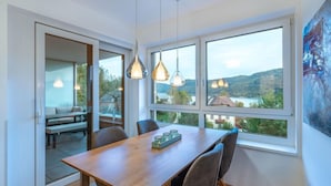 A dining area with a wooden table and chairs, large windows with a view of a lake and hills, pendant lights, and a sliding glass door leading to an outdoor seating area. - Seeblick alpe maritima Ski & See-Top 12 by Interhome (Ossiacher See)