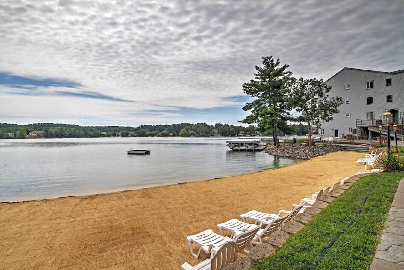 A lakeside view with a sandy beach, lounge chairs, a dock, and a building with a balcony.