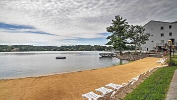 A lakeside view with a sandy beach, lounge chairs, a dock, and a building with a balcony.