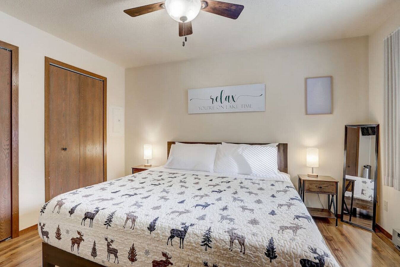 Une chambre à coucher avec un lit, un ventilateur de plafond, une table de chevet avec une lampe, un miroir et un panneau accroché au mur.