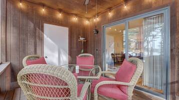 Un área de estar techada al aire libre con sillas de mimbre y una mesa de vidrio, paredes de madera y un ventanal corredero que da a un espacio interior.