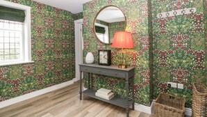 Un pasillo con papel tapiz floral, un espejo redondo, una mesa con un reloj, una lámpara y una cesta de mimbre.