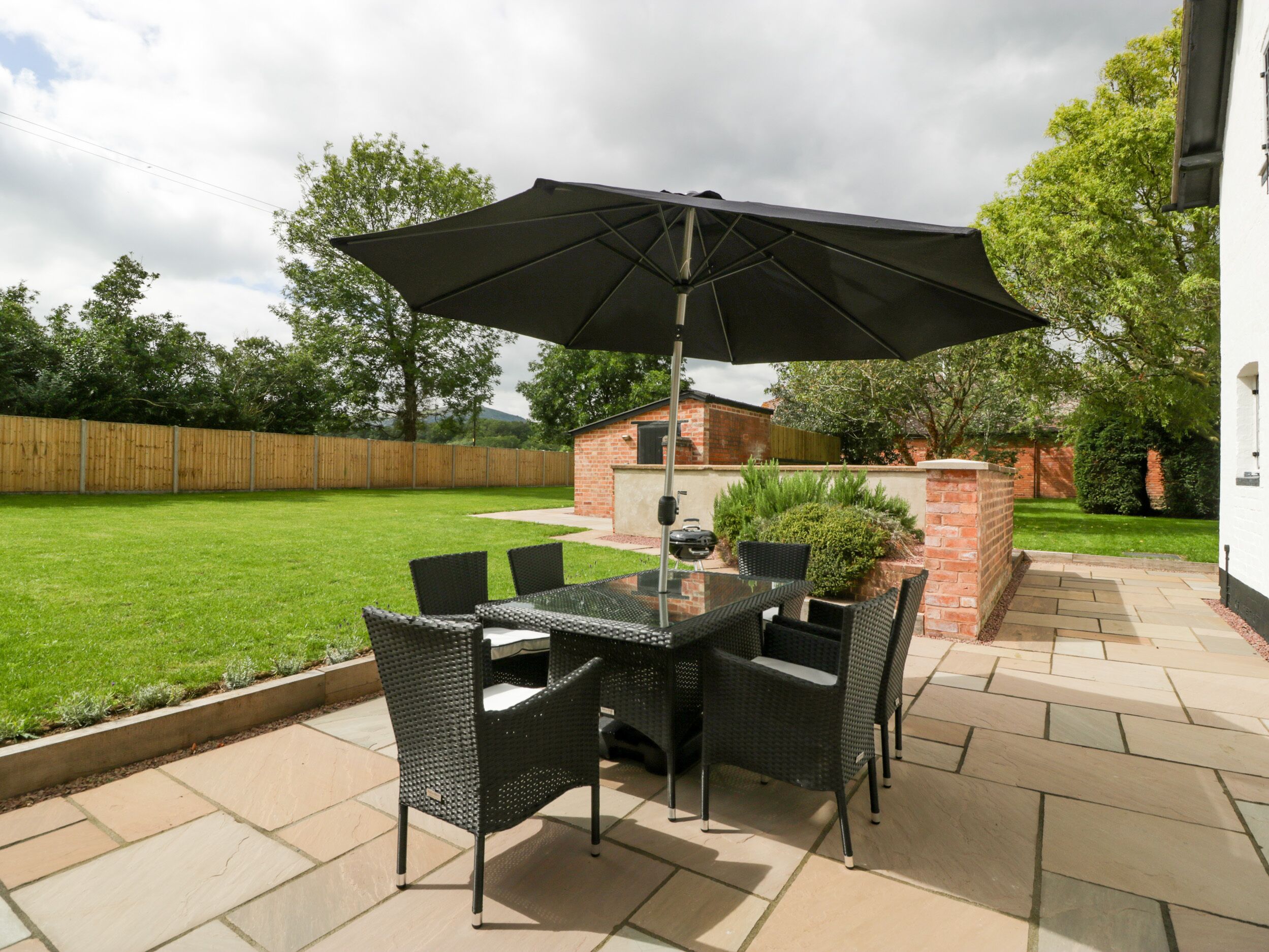 An outdoor patio with a glass table and chairs, a large umbrella, and a barbecue grill.