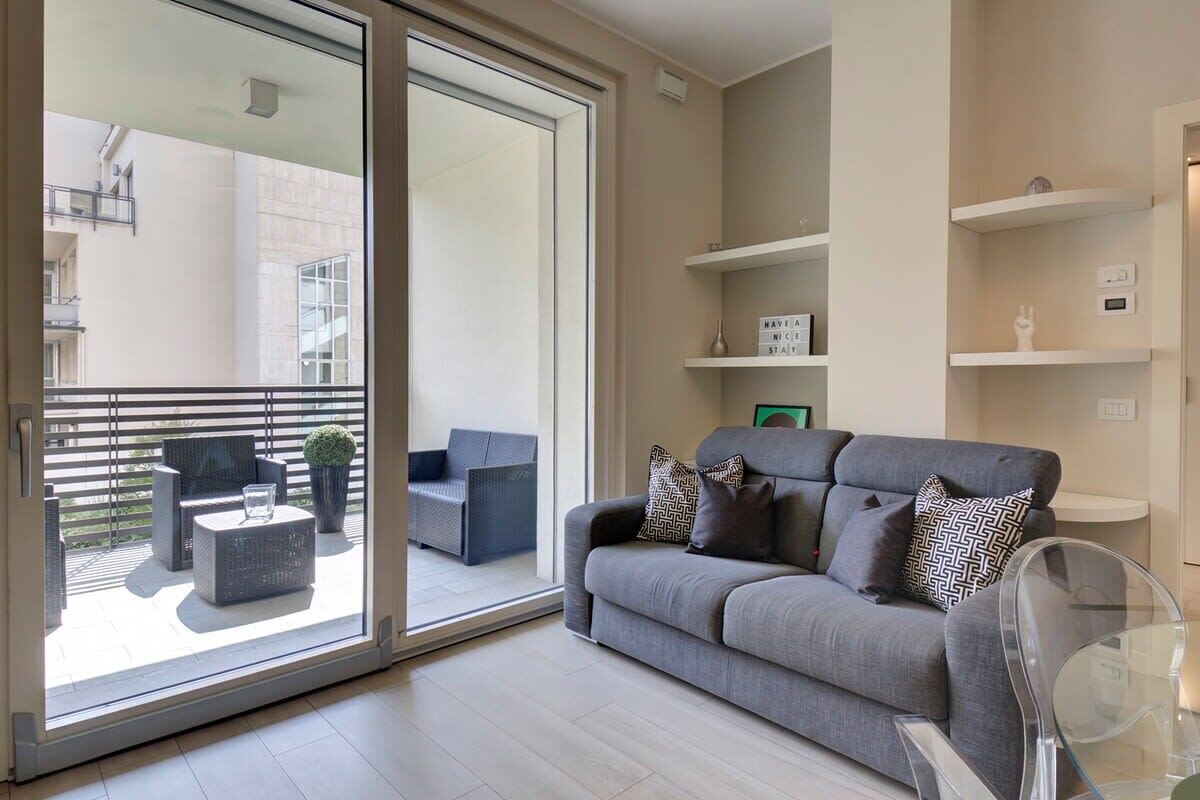 Uma sala de estar moderna com um sofá cinzento, uma mesa de centro de vidro transparente e uma varanda com mobiliário exterior.