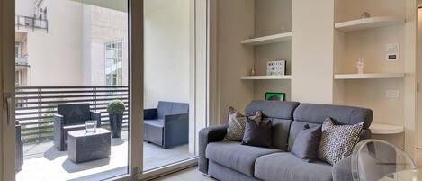 Uma sala de estar moderna com um sofá cinzento, uma mesa de centro de vidro transparente e uma varanda com mobiliário exterior.