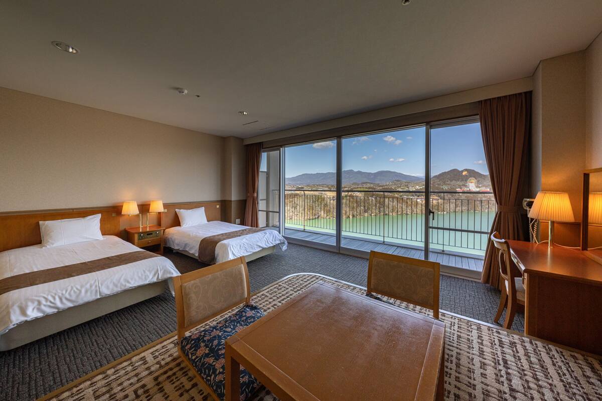 Habitación de hotel con dos camas, una mesa de comedor y un ventanal con vistas a montañas y un campo de golf.