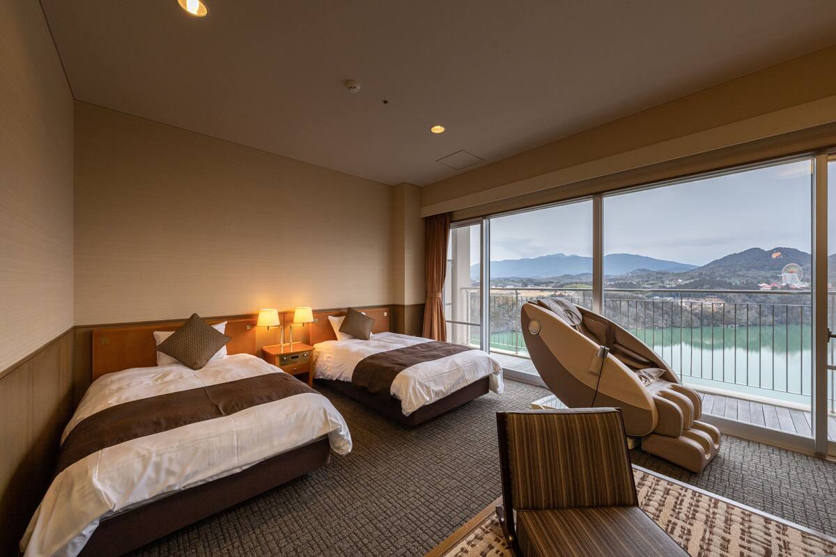 Habitación de hotel con dos camas, una silla de masajes y un ventanal amplio con vistas a un cuerpo de agua y montañas.