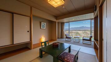 A traditional Japanese room with tatami flooring, a low table with a cushion, a sliding door, and a view of mountains through large windows.