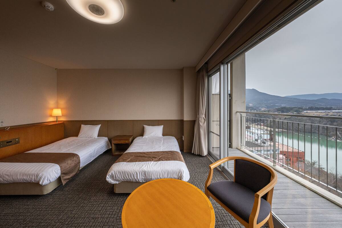 Habitación de hotel con dos camas individuales, un sofá, una mesita y un balcón con vistas a un río y montañas.