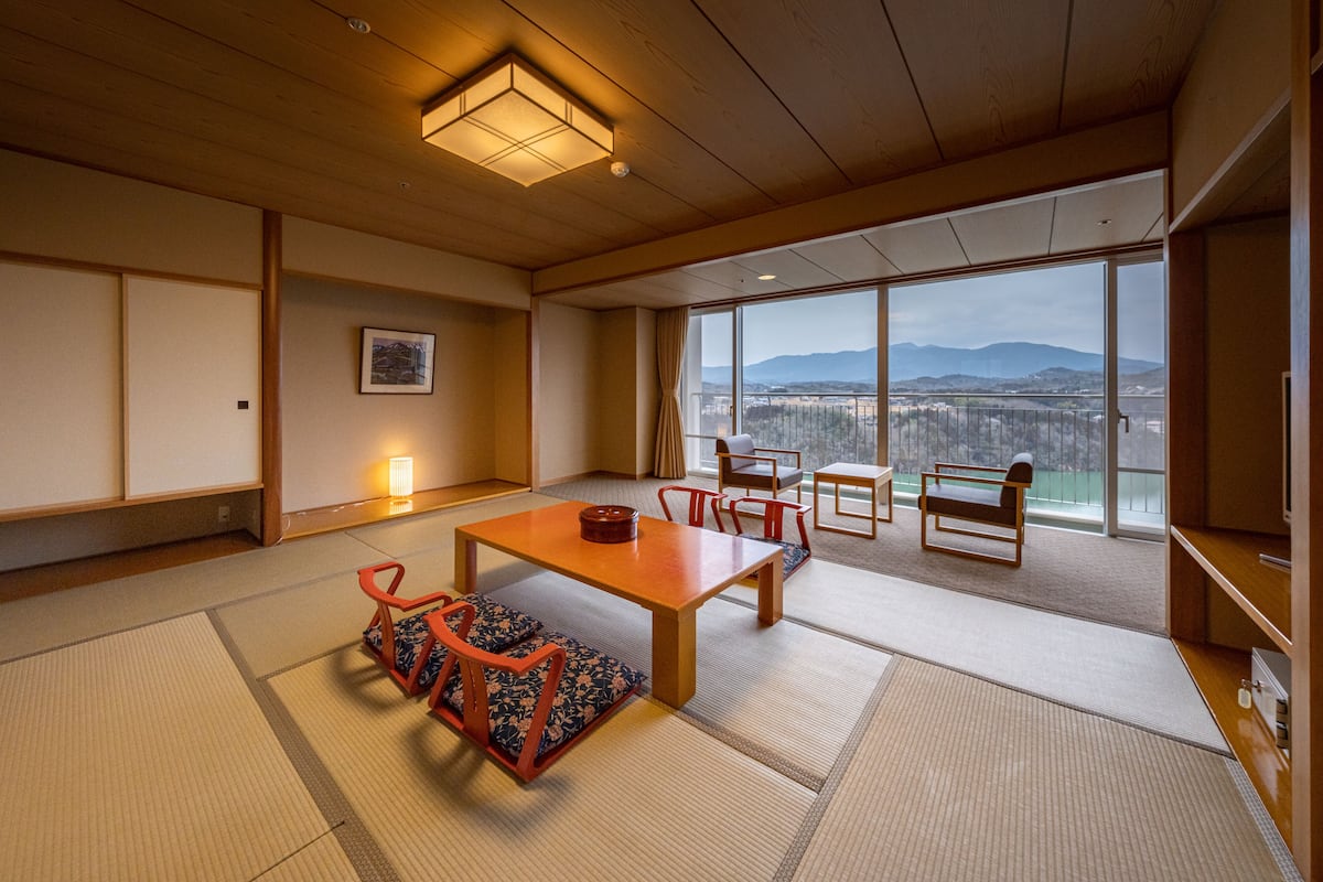 Una habitación japonesa tradicional con suelo de tatami, una mesa de madera baja y vistas a un paisaje urbano a través de amplios ventanales.