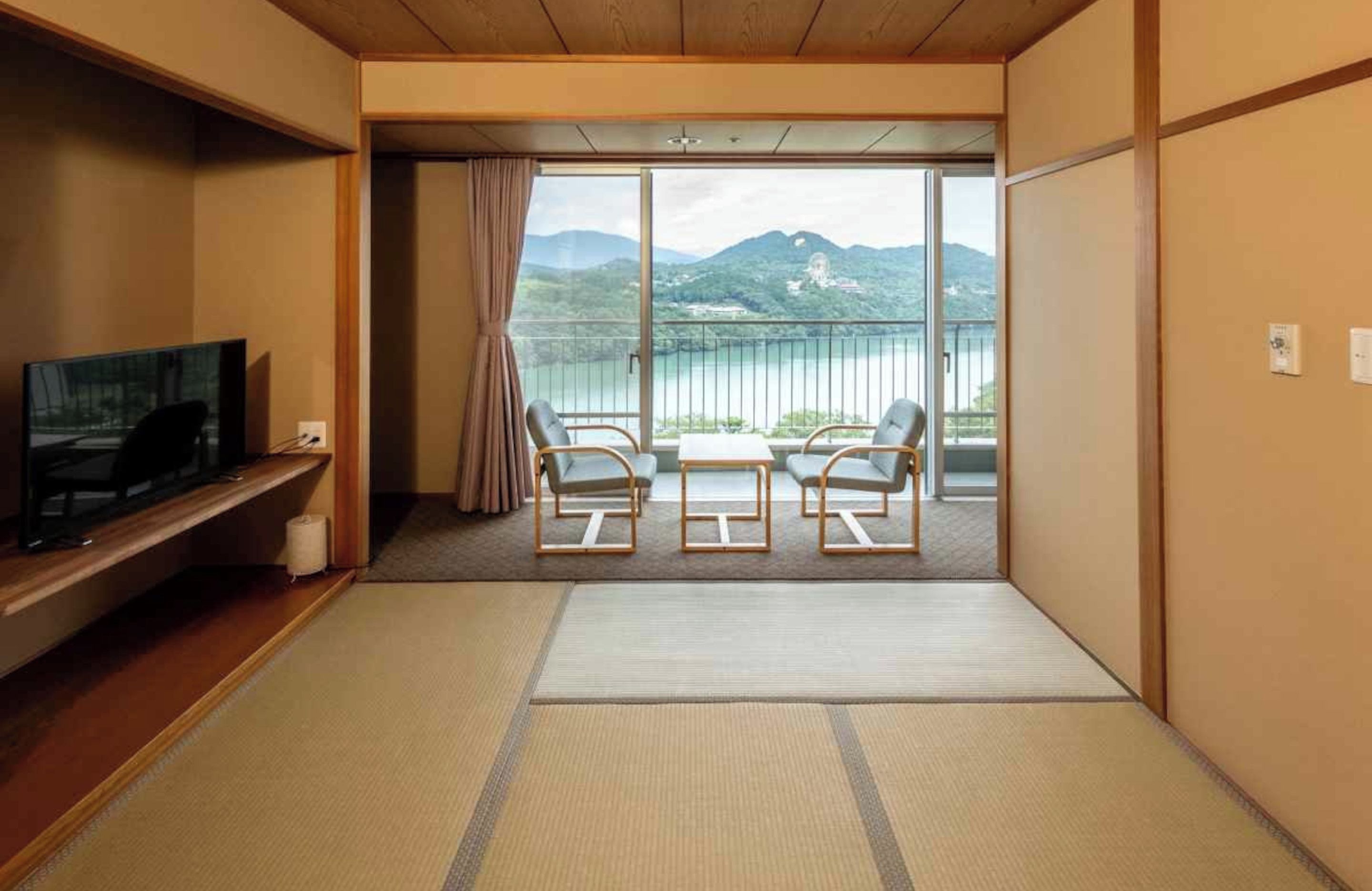 A hotel room with a television, a sofa, two chairs, a sliding glass door leading to a balcony with a view of mountains and a lake.
