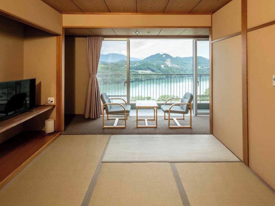 A hotel room with a television, a sofa, two chairs, a sliding glass door leading to a balcony with a view of mountains and a lake.