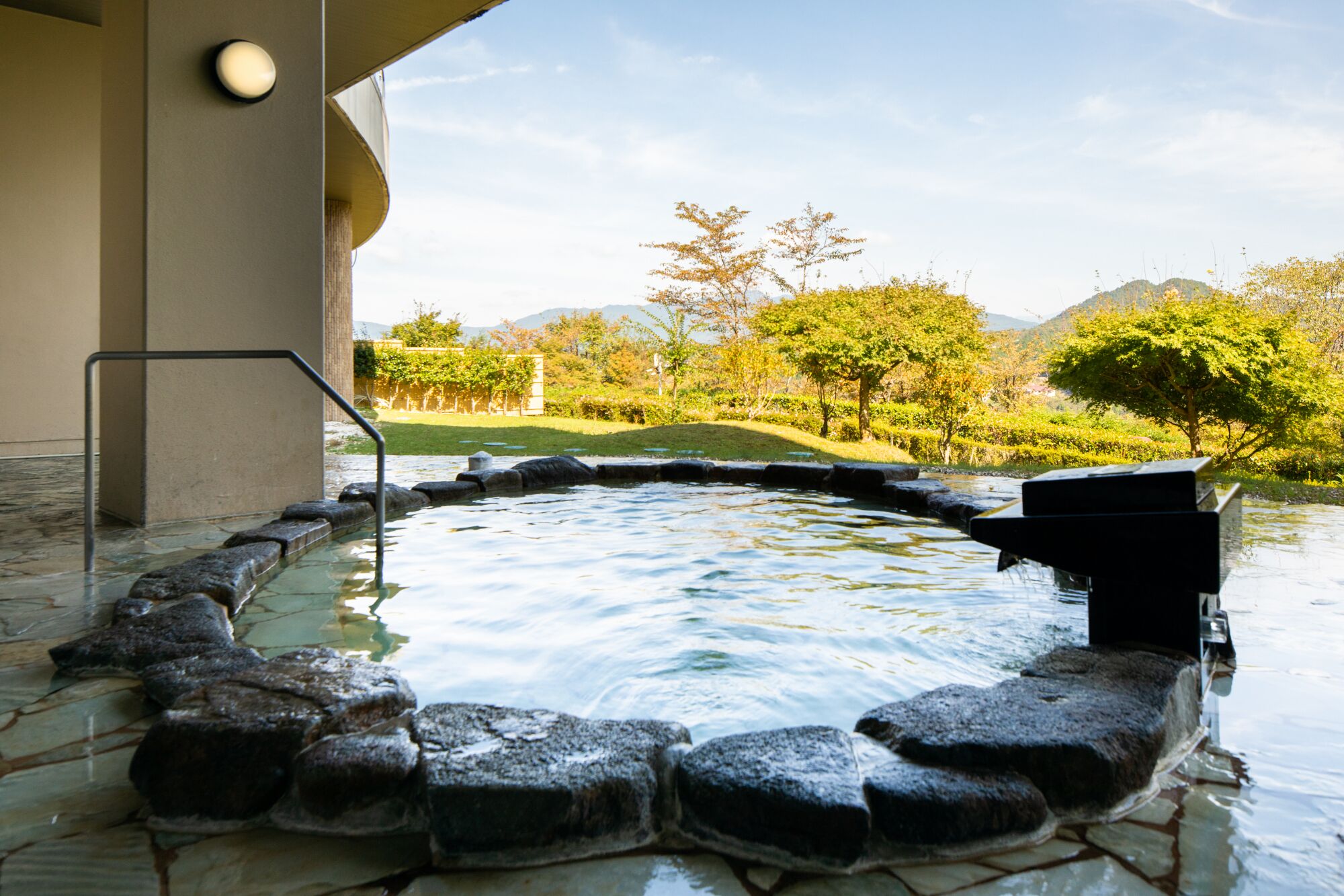 A hot tub with stone steps, surrounded by a stone wall, with a view of a landscaped garden and trees in the background.
