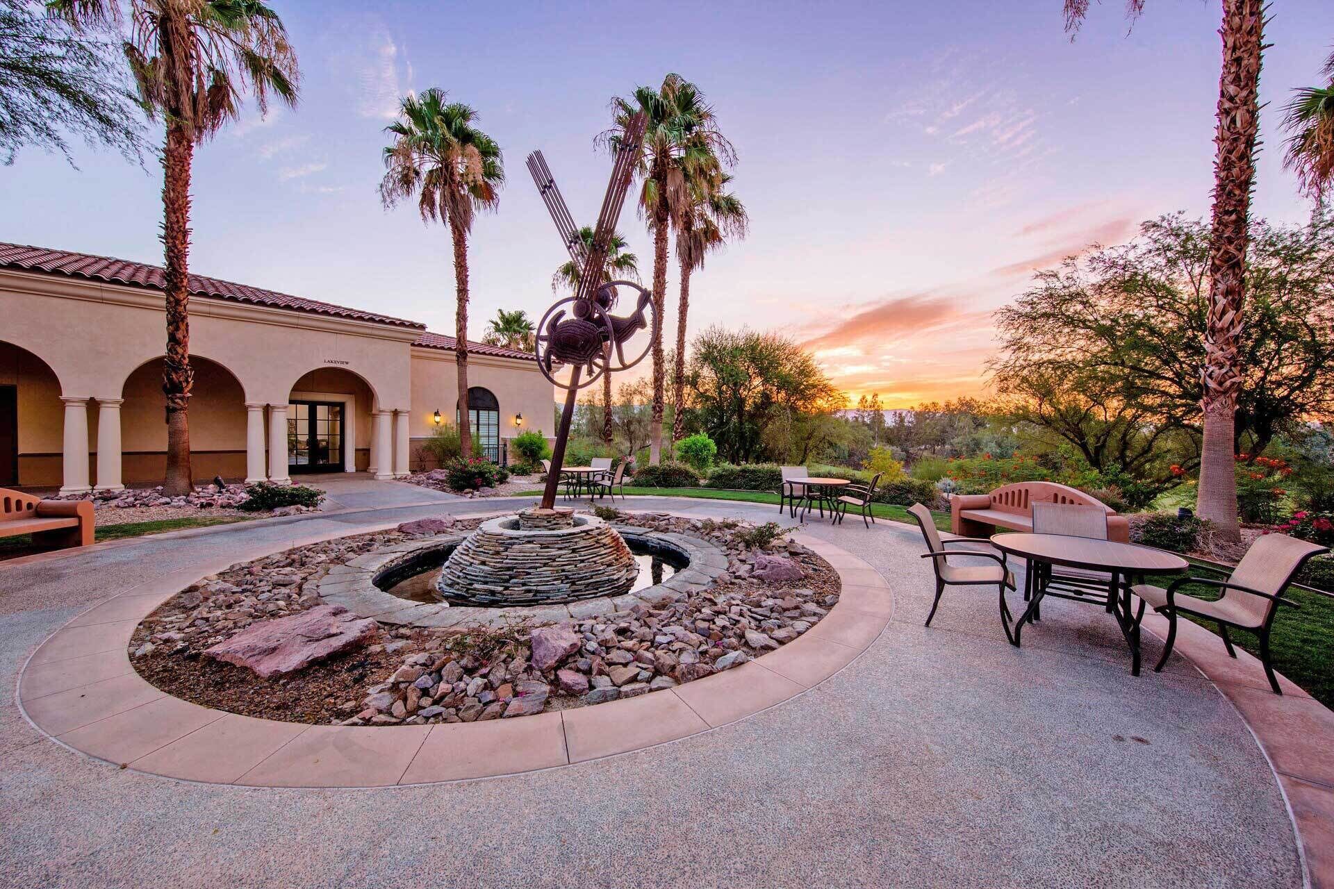 A courtyard with a circular stone feature, outdoor seating, palm trees, and a building with arched doorways.