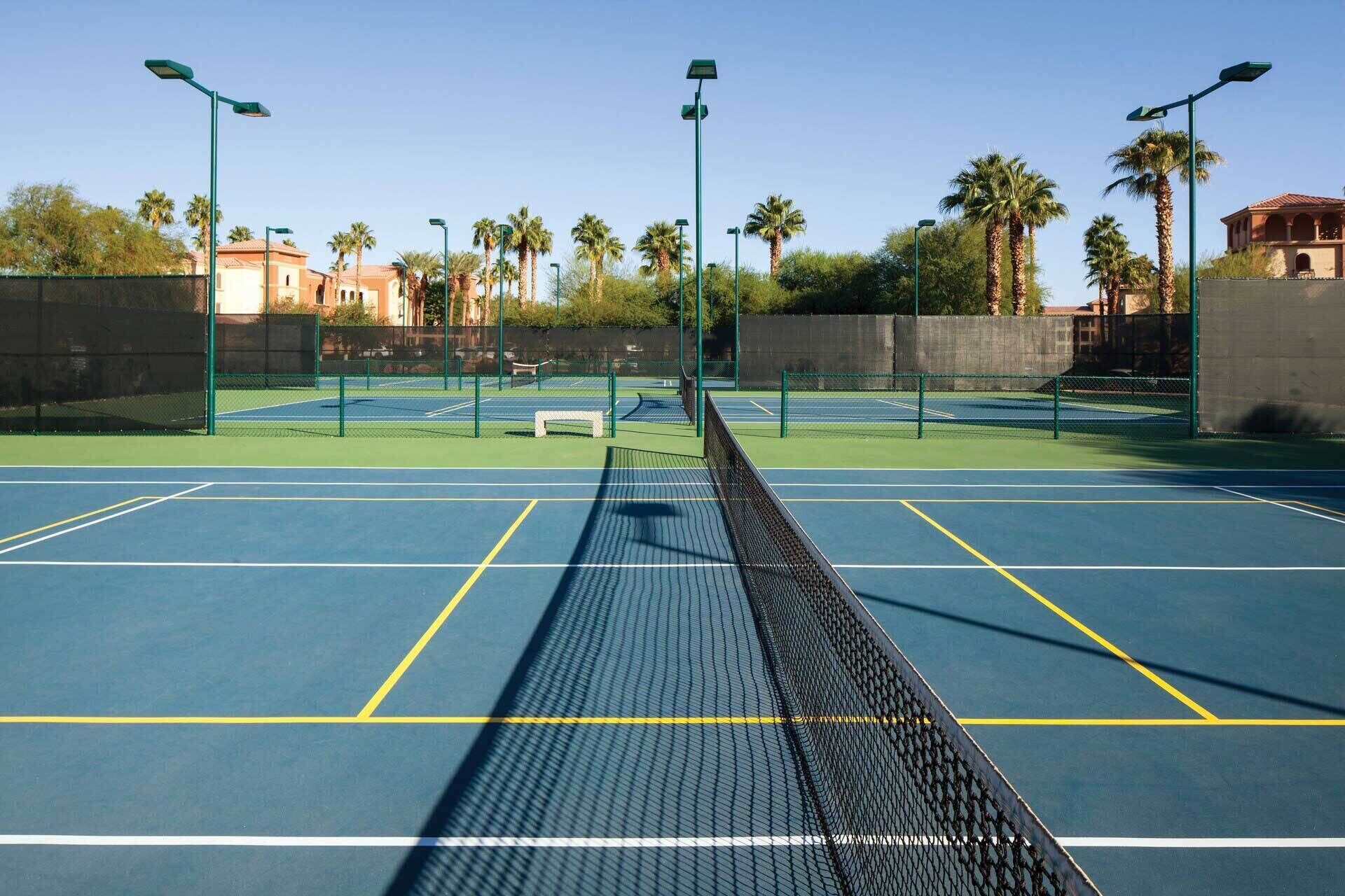 Una cancha de tenis al aire libre con iluminación verde, rodeada de palmeras y una valla.