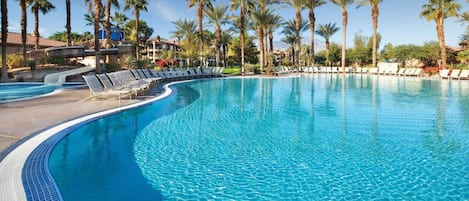 Une grande piscine à eau claire et bleue, de forme incurvée, entourée de palmiers et de chaises longues.
