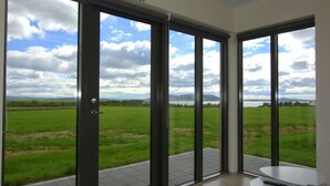 A room with large sliding glass doors leading to a grassy field and a lake, with a small table and chair inside. - Cottages built for privacy and unobstructed views (Bláskógabyggð)