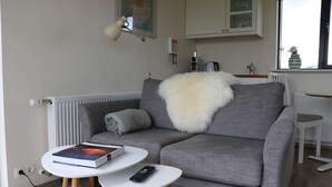 A living room with a grey sofa, a white fluffy throw, a remote control, and a book on a small white table. - Cottages built for privacy and unobstructed views (Bláskógabyggð)