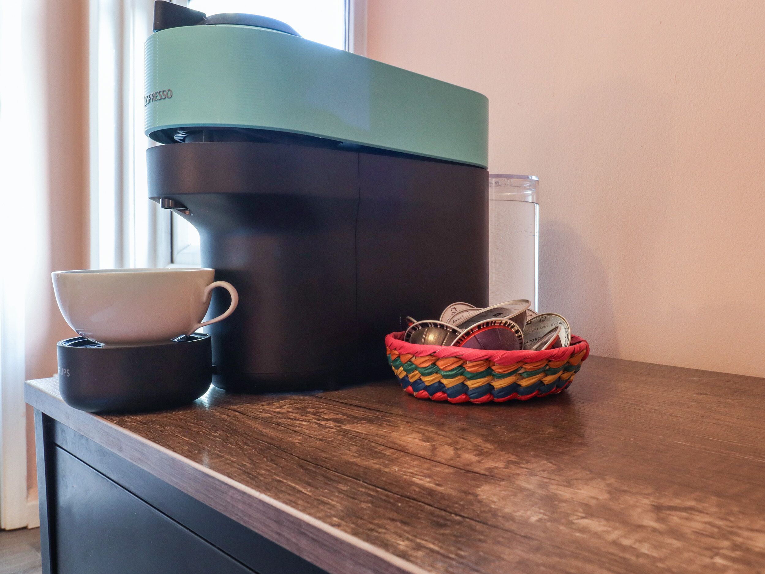 Een koffiezetapparaat, een kop en een mandje muntstukken op een houten tafel.