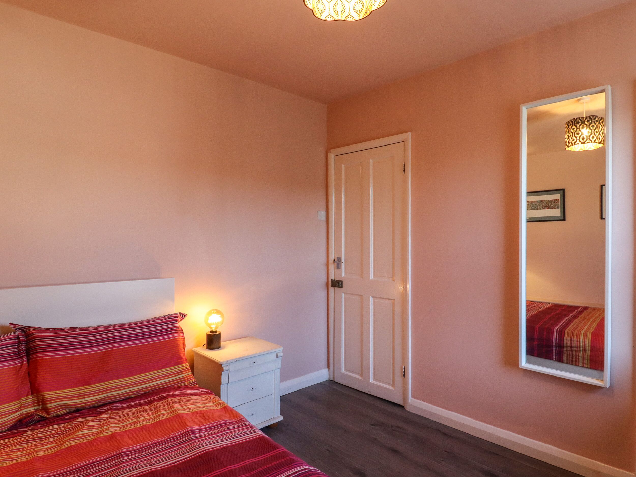 A bedroom with a bed, a nightstand with a lamp, a door, and a mirror on the wall.