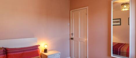A bedroom with a bed, a nightstand with a lamp, a door, and a mirror on the wall.