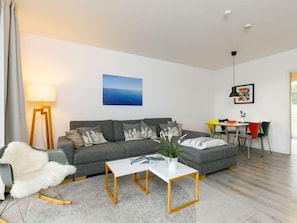 Ein modernes Wohnzimmer mit einem grauen Sofa, einem weißen Couchtisch und einem gemütlichen Sessel. Es gibt einen Essbereich mit Tisch und Stühlen, und eine Lampe steht in der Ecke.