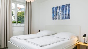 Une chambre à coucher moderne avec un lit blanc, une table de chevet, une lampe et une fenêtre avec des rideaux.
