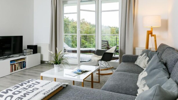 Un salon moderne avec un canapé gris, une table basse blanche, un téléviseur posé sur un meuble blanc, une bibliothèque et un balcon offrant une vue sur la verdure.