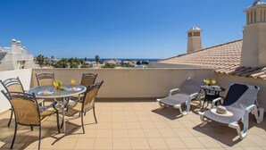 A rooftop terrace with a table and chairs, a small table with drinks, and a view of the sea and buildings.