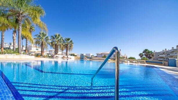 A clear blue swimming pool with a metal ladder, surrounded by palm trees and modern buildings.