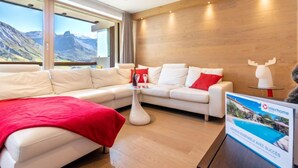 A modern hotel room with a white sofa, red cushions, a glass coffee table, and a view of mountains through large windows.