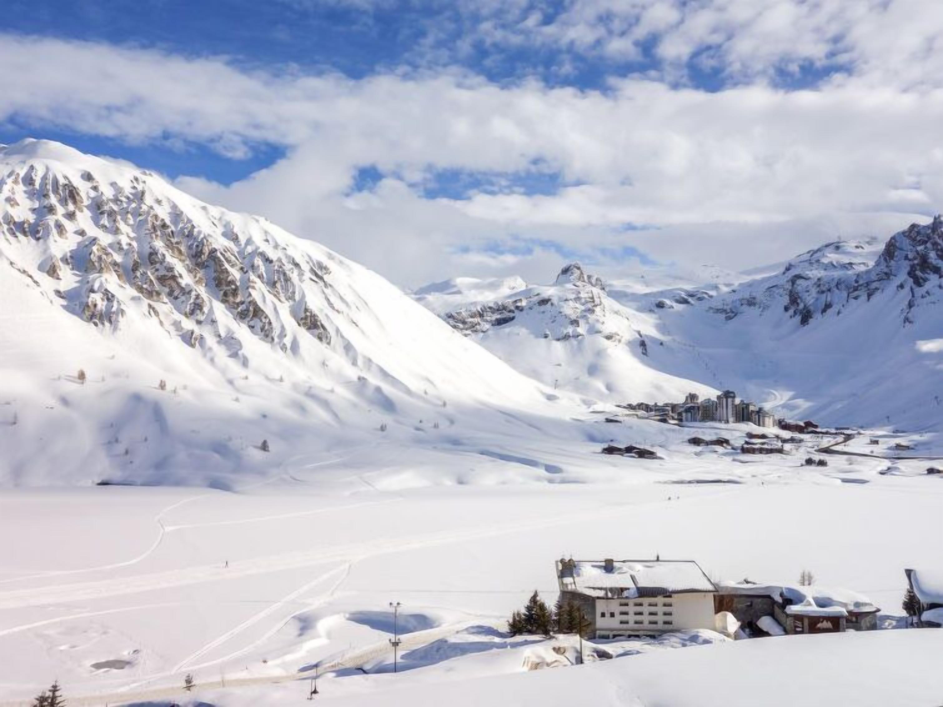 Un paysage montagneux enneigé, avec un petit ensemble de bâtiments au premier plan.