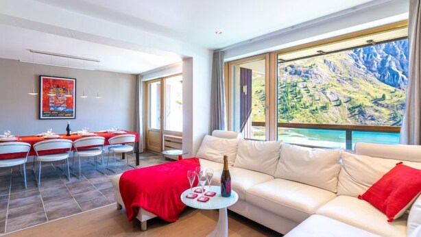 A modern hotel room with a large window offering a mountain view, a dining area with red tablecloths, and a white sofa with red cushions.