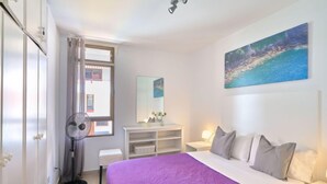Une chambre à coucher comprenant un lit, une table de chevet, une commode, une fenêtre avec des rideaux, un ventilateur de plafond et un tableau accroché au mur.