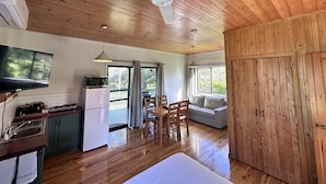 Interior em madeira com ventilador de teto, TV na parede, cozinha compacta, área de jantar com mesa e cadeiras, e um sofá.
