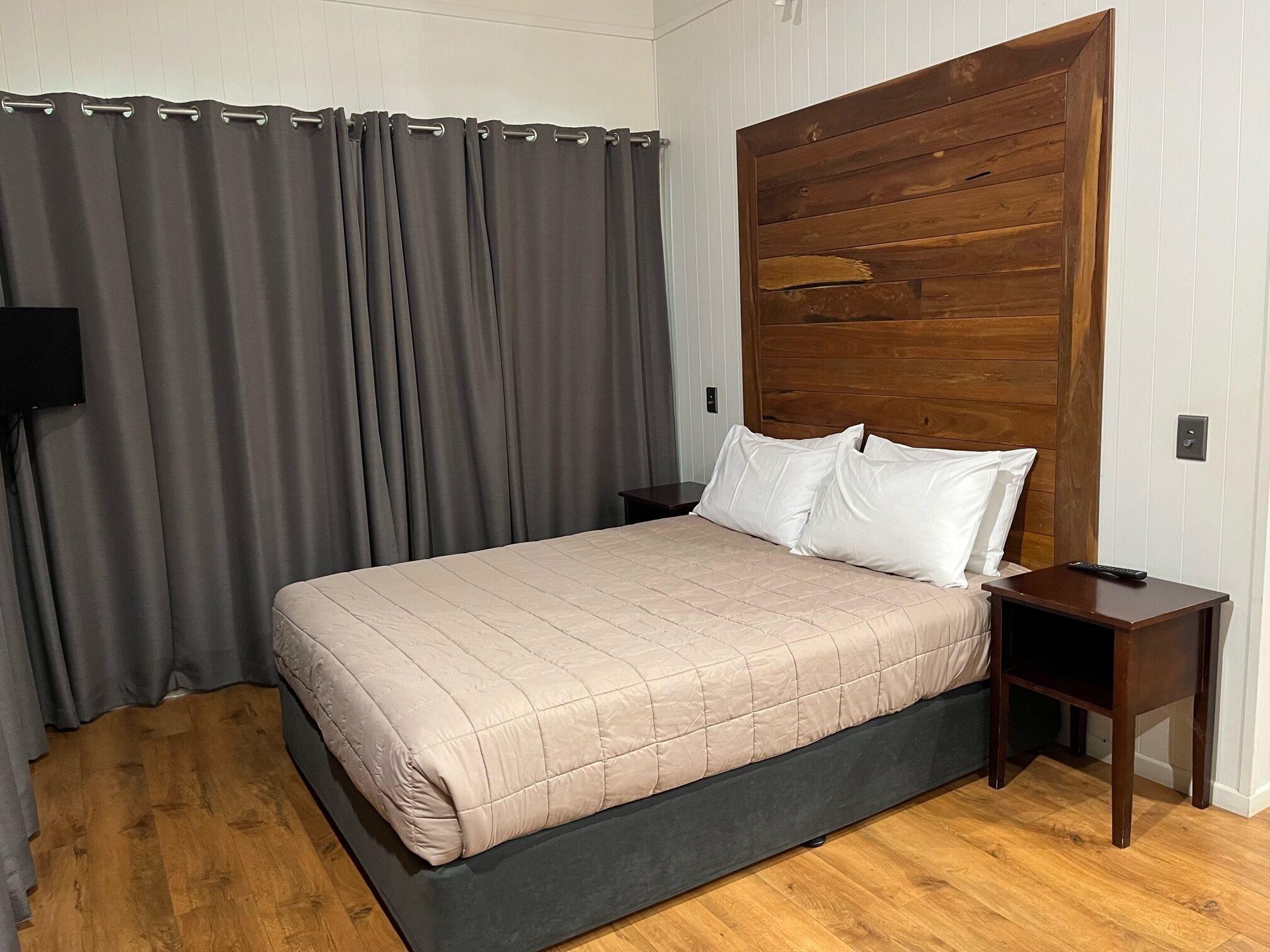 A bedroom with a bed, a wooden headboard, a nightstand, a remote control, and a grey curtain.