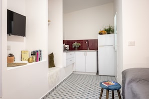 A compact room with a kitchenette, a bed, a small stool, and a shelf with books and a globe.