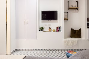 A room with a white wardrobe, a mounted TV, a shelf with books and decorative items, and a patterned floor.